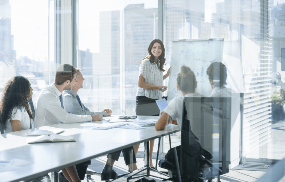 A woman stands in front of a flipchart as she presents to her colleagues in a meeting room.